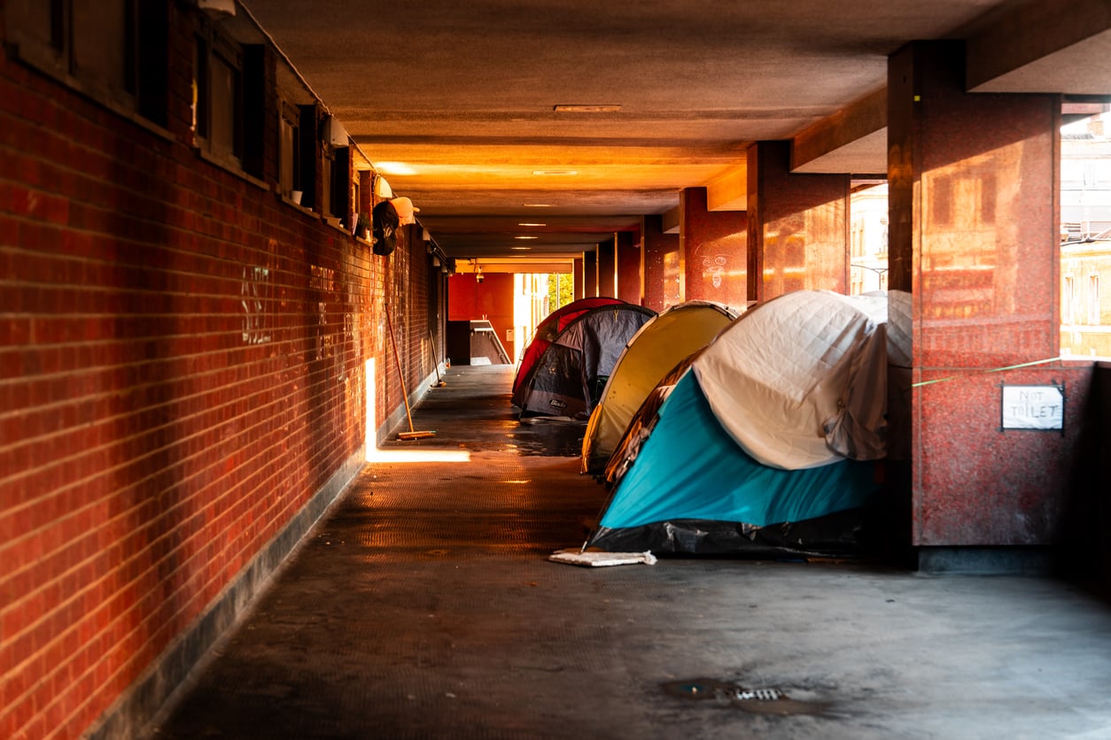 Several tents are set up along the wall of an urban covered walkway with red brick and pillars, a quiet testament to life in 2026 problems. Sunlight shines through, highlighting the tents. A small sign reading “toilet” is visible on the right.