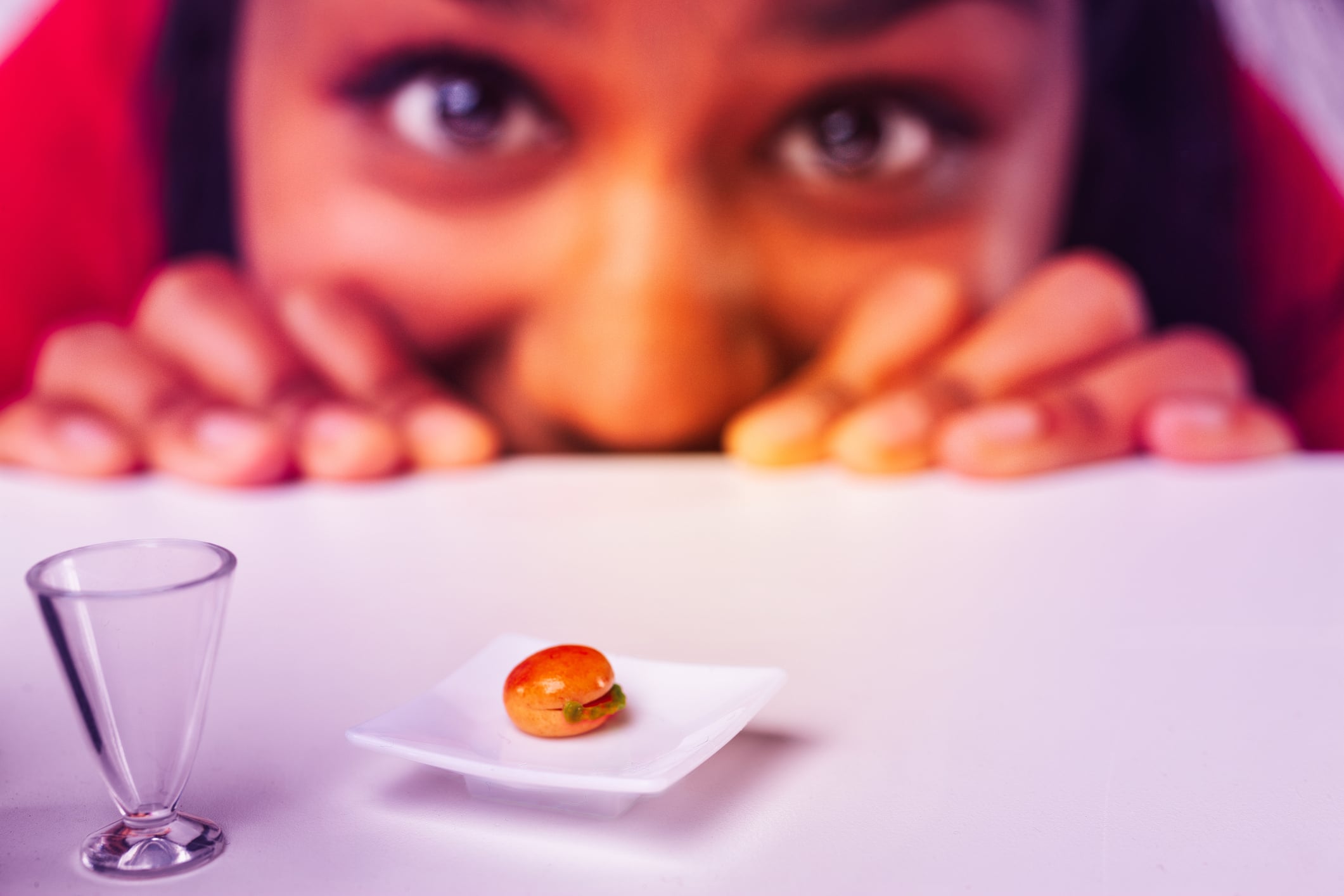 A person eagerly looks at a tiny burger on a small white plate, next to a miniature glass, all placed on a white surface.
