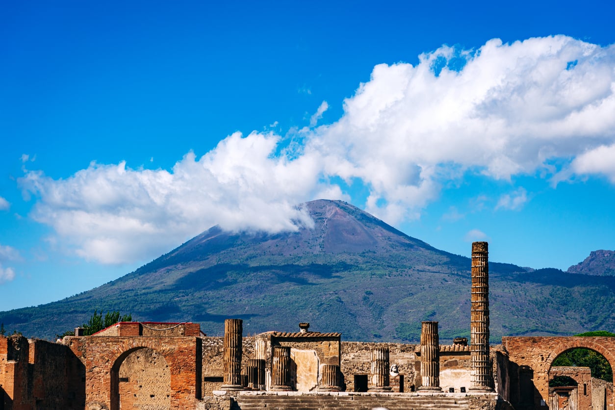 Ancient ruins with stone columns and walls are set in front of a green mountain under a bright blue sky with scattered clouds.