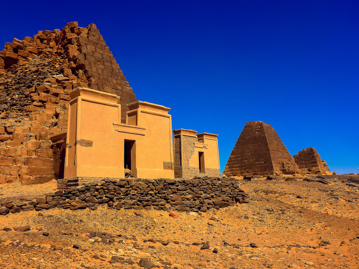 Ancient Nubian pyramids and tombs rise from a sandy desert under a clear, deep blue sky. The weathered pyramids, with some partially crumbled structures visible, stand as striking reminders of history—even as the region appears on recent lists of Places Travelers Are Warning Against Going In 2026.