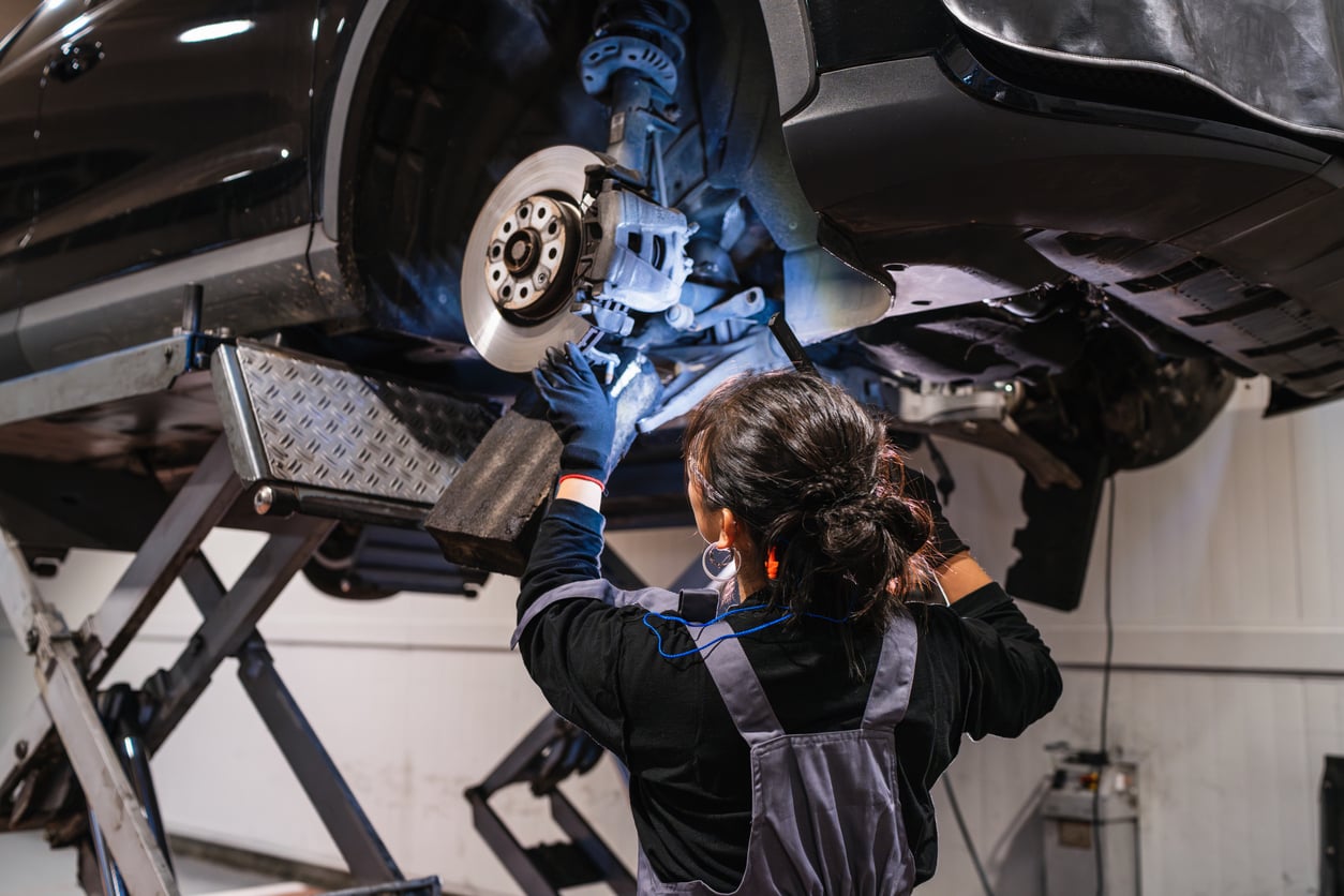 A mechanic wearing gloves and overalls works on the exposed brake system of a car lifted on a hydraulic lift in a workshop.