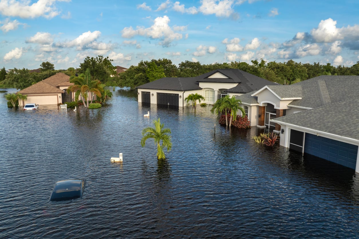 Flooded suburban neighborhood with several one-story houses partially underwater, palm trees standing in water, and a mostly submerged car. The sky is blue with scattered clouds.