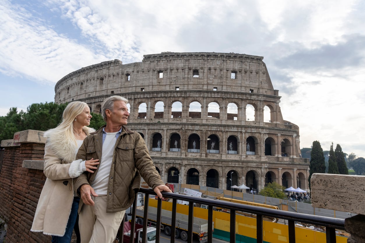 A couple stands near a railing, smiling and looking away, with the Colosseum in Rome in the background under a partly cloudy sky—a timeless scene at one of the travel destinations boomers are skipping.