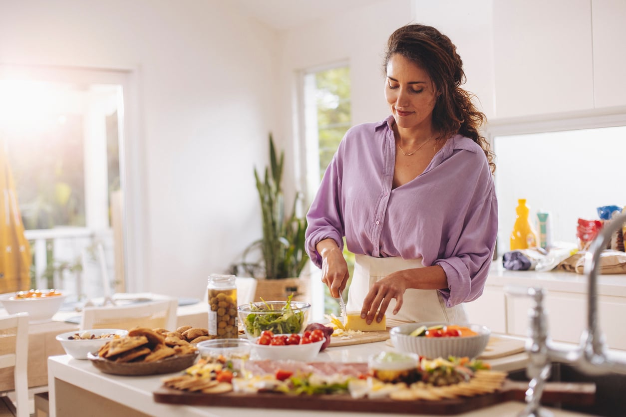 A woman in a lavender shirt chops vegetables in a bright kitchen, surrounded by bowls of salad, snacks, and a platter of appetizers on the counter. Sunlight streams through the windows.