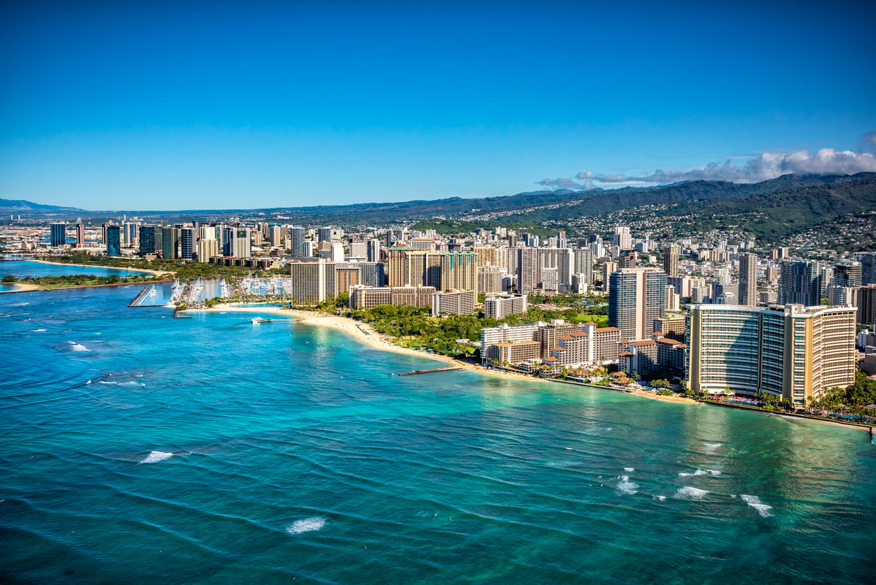 Aerial view of a city skyline with tall buildings along a sandy beach, turquoise ocean water, and mountains in the background under a clear blue sky—an idyllic setting that offers everything from breathtaking scenery to vibrant nightlife, though some visitors have noted the challenge of finding anything beyond bad food in certain tourist areas.