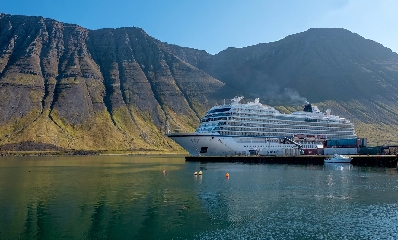 A white cruise ship is docked at a pier, surrounded by calm water with mountain cliffs in the background under a clear blue sky.