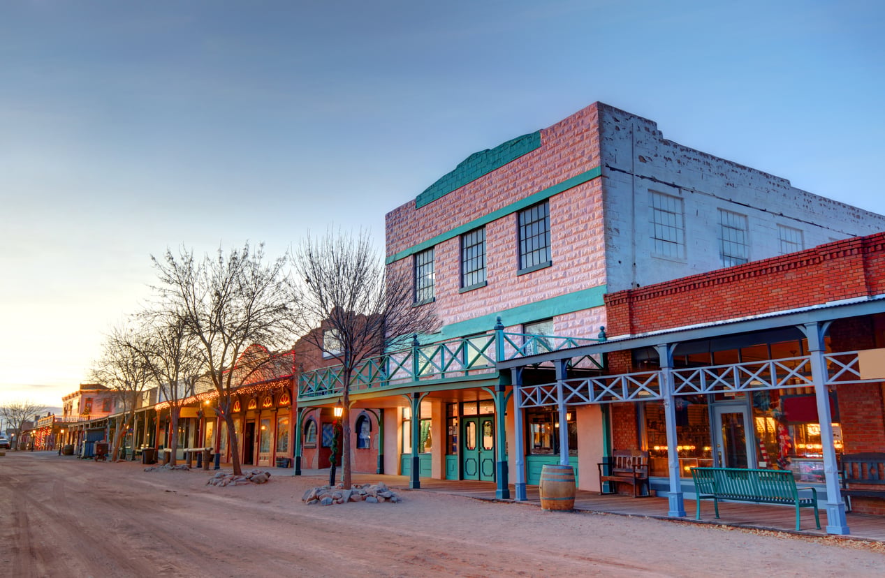 Colorful historic buildings line a dusty, empty street in a Western-style old town at sunset, with wooden benches, barrels, and bare trees visible—evoking the charm of nostalgic travel destinations.