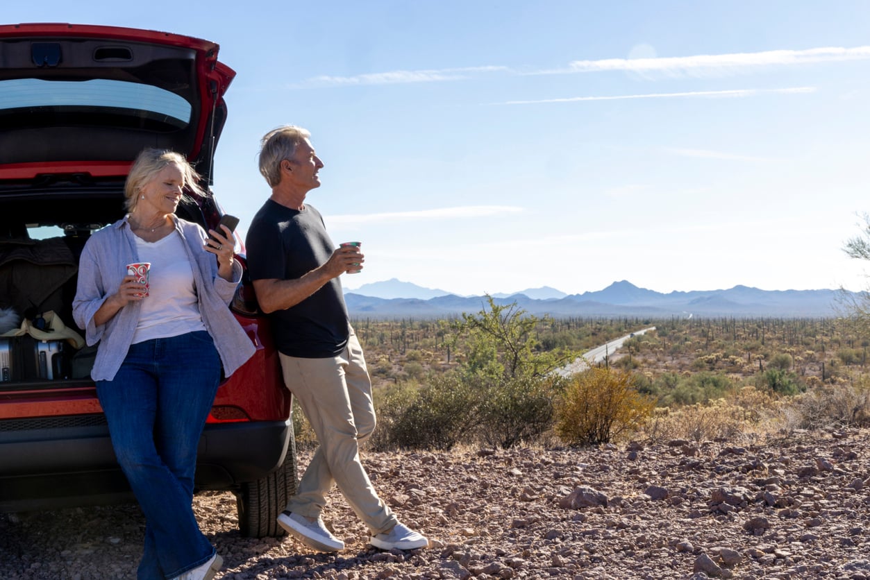 An older couple stands by an open car trunk in a desert landscape, holding drinks and smiling. Mountains and a winding road are visible in the background under a clear blue sky.