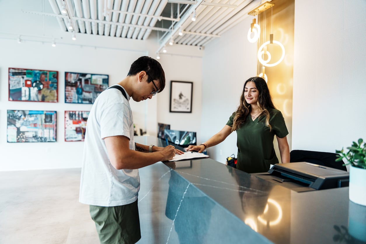 A person signs a form at a modern reception desk while a receptionist smiles and assists them. Framed art hangs on the white wall in the background.