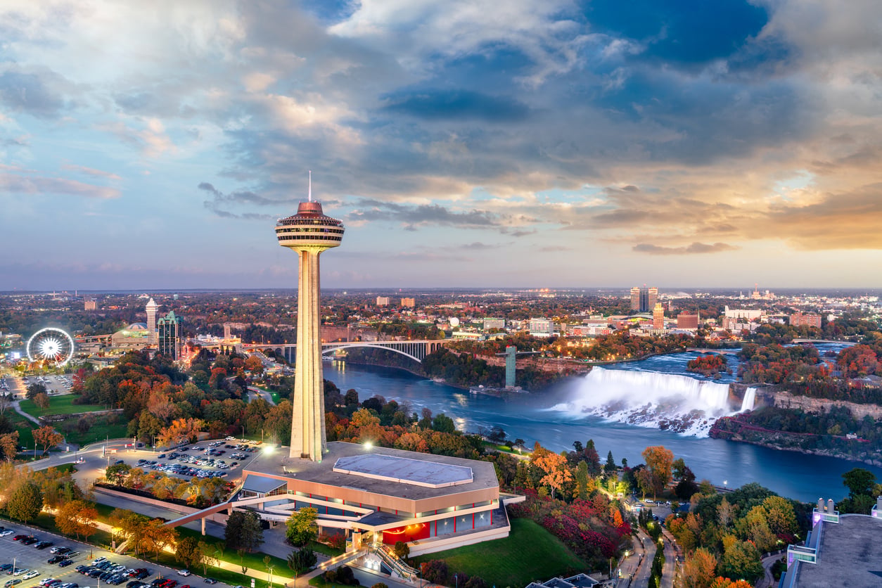 A cityscape of Niagara Falls with the Skylon Tower in the foreground, waterfalls in the middle distance, and a sunset sky with colorful clouds above—despite the breathtaking view, visitors sometimes lament the bad food options nearby.