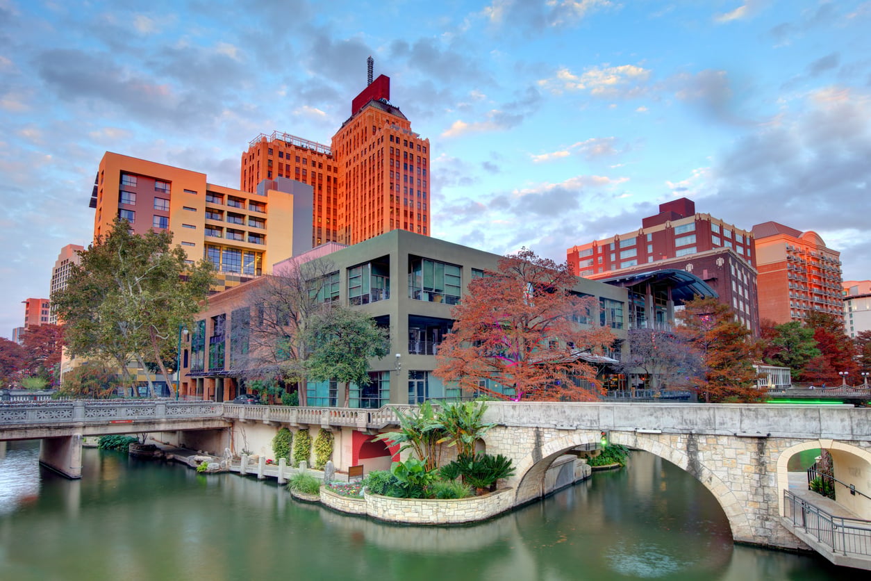 Downtown San Antonio’s River Walk, one of the city’s most beloved vacation spots, features a stone bridge over a calm canal, colorful fall trees, and tall buildings in the background under a partly cloudy sky at sunset.