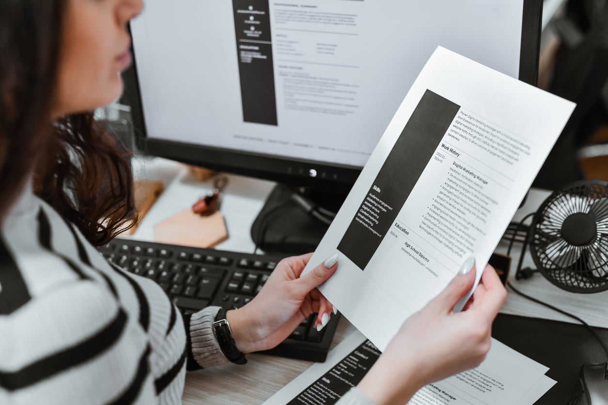 A person reviews printed resumes at a desk with a computer displaying a digital resume, keyboard, and a small fan visible in the background.