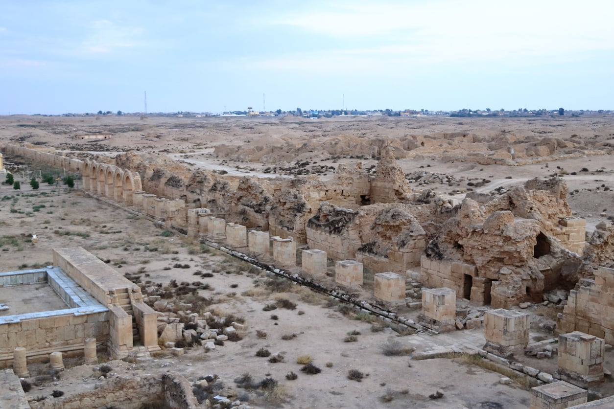 Ancient stone ruins stretch across a desert landscape under a cloudy sky, with scattered vegetation and distant buildings visible on the horizon—a haunting scene from one of the places travelers are warning against going in 2026.
