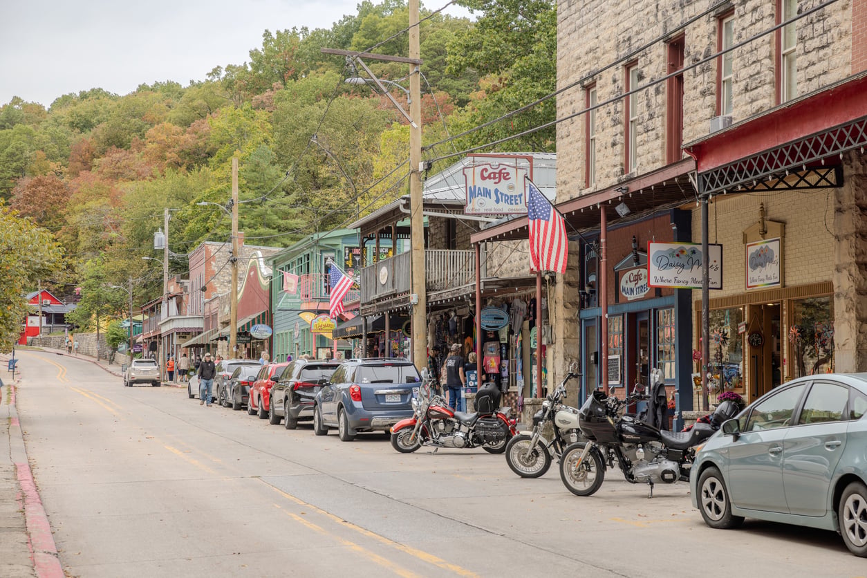 A small-town street lined with shops, parked cars, and motorcycles—one of the best fall towns in America. American flags hang from buildings, and trees with autumn leaves fill the background. People walk along the sidewalk under cloudy skies.