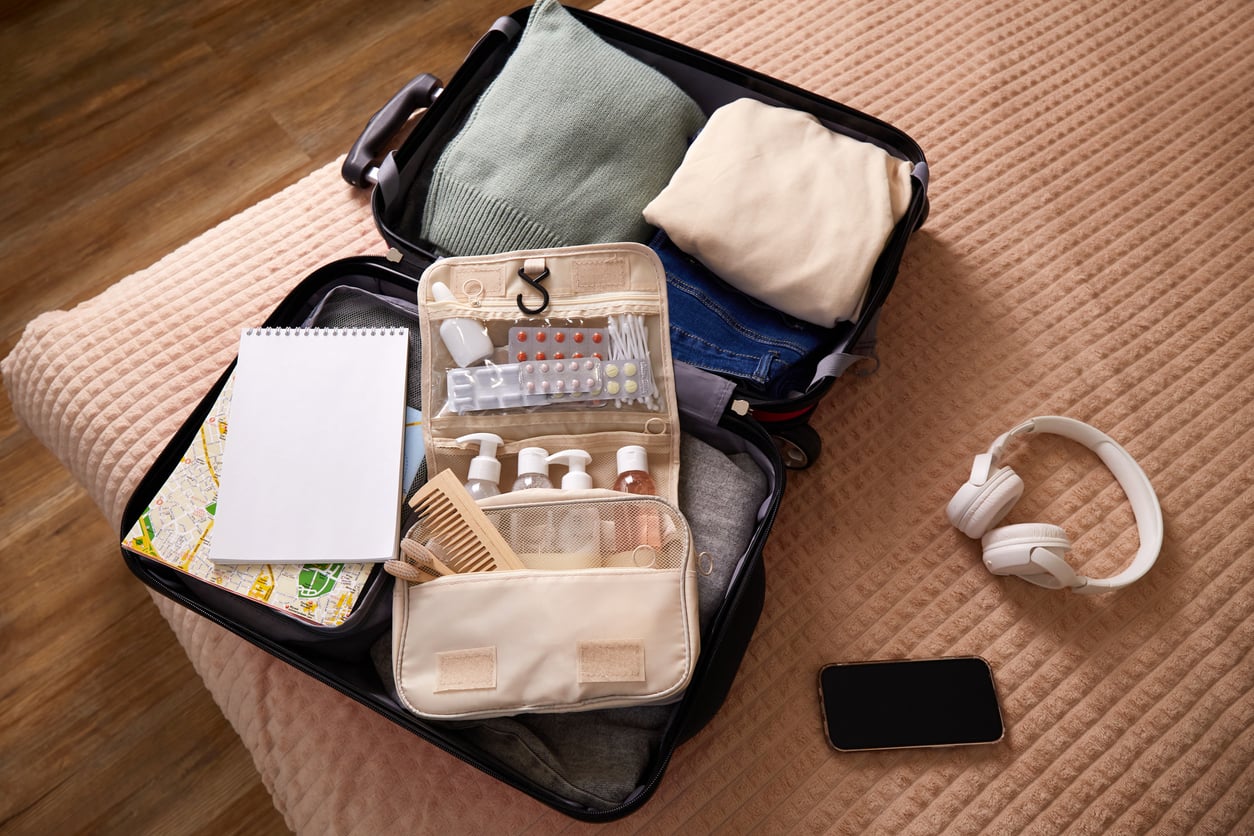 Open suitcase on a bed with neatly packed clothes, toiletries, a map, notepad, comb, and blister packs of pills. Nearby are white headphones and a smartphone on a peach-colored quilted blanket.
