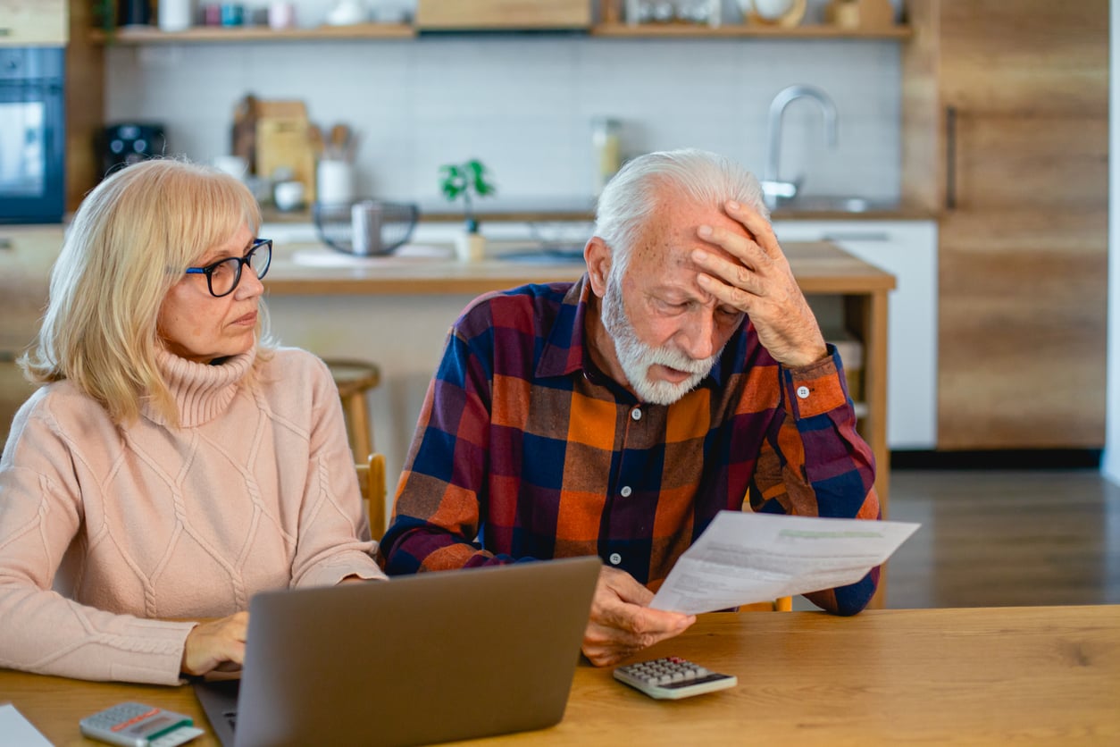 An elderly couple sits at a table with a laptop and papers. The man looks worried, holding his forehead and a document, while the woman sits beside him, looking at the paper with concern.