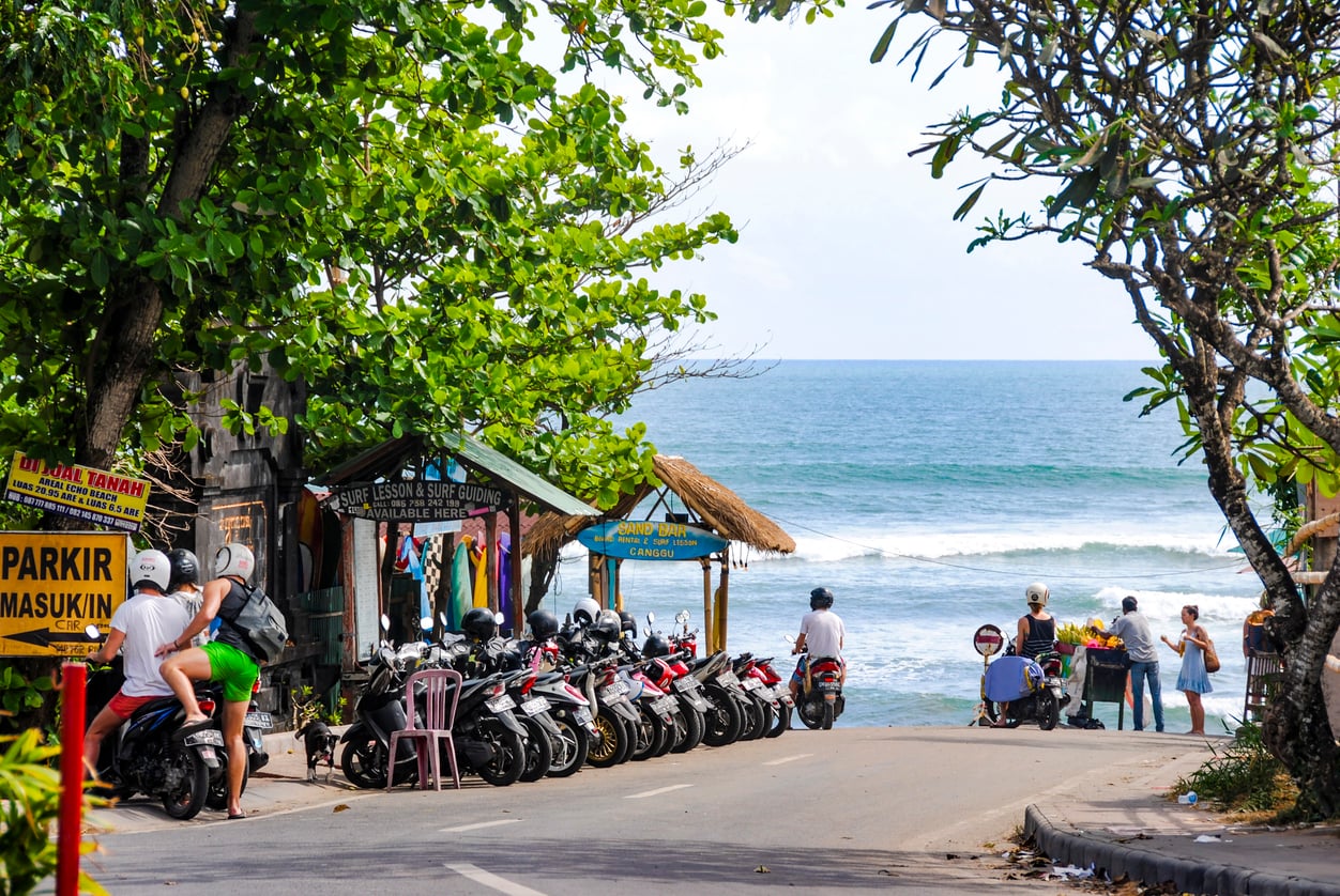 Motorbikes parked near a beachside road with people gathered by the ocean, trees providing shade, and small huts selling clothes and goods.