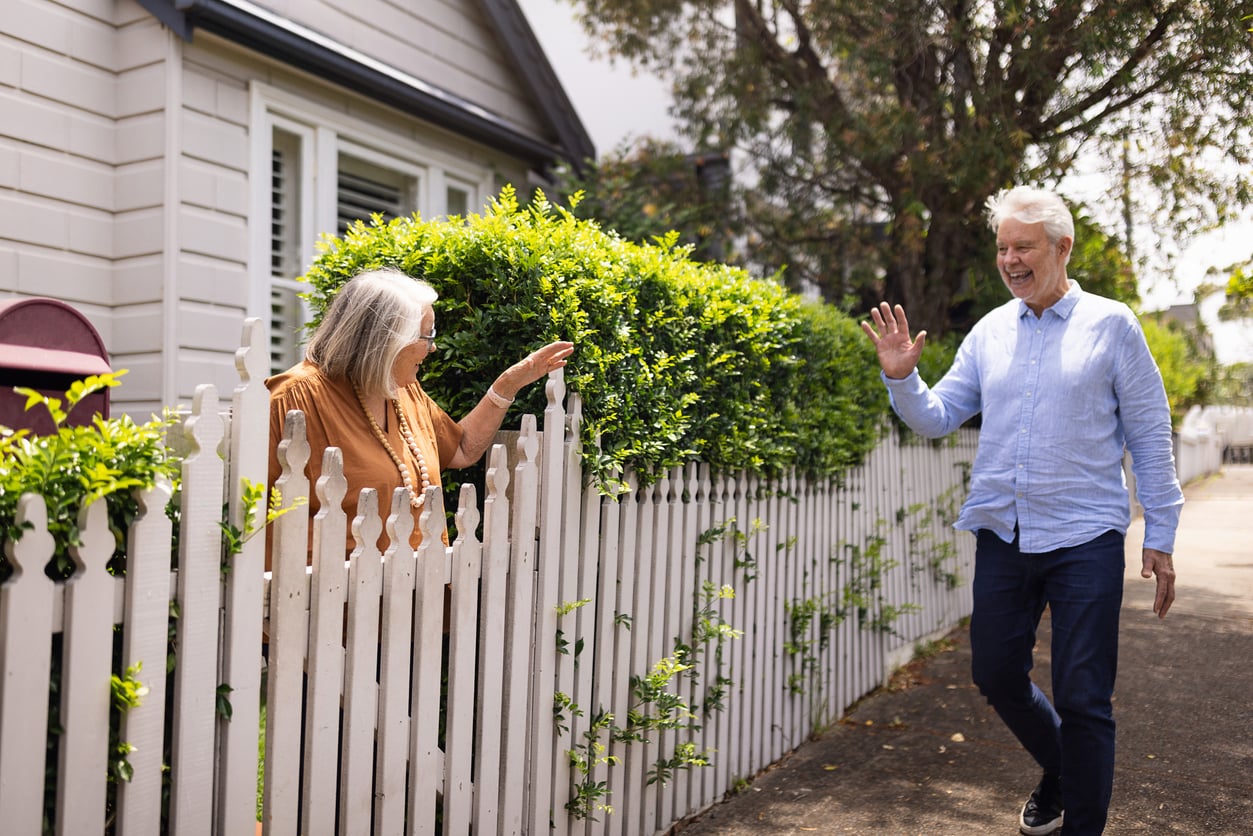 An older woman stands by a white picket fence, smiling and waving to an older man walking on the sidewalk. The scene is bright and friendly, with greenery and a white house in the background.