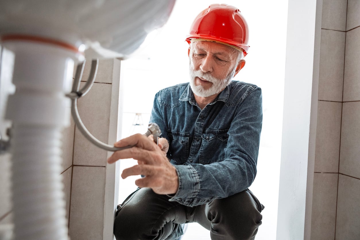 An older man wearing a red hard hat and denim shirt kneels and repairs plumbing under a sink with a wrench.