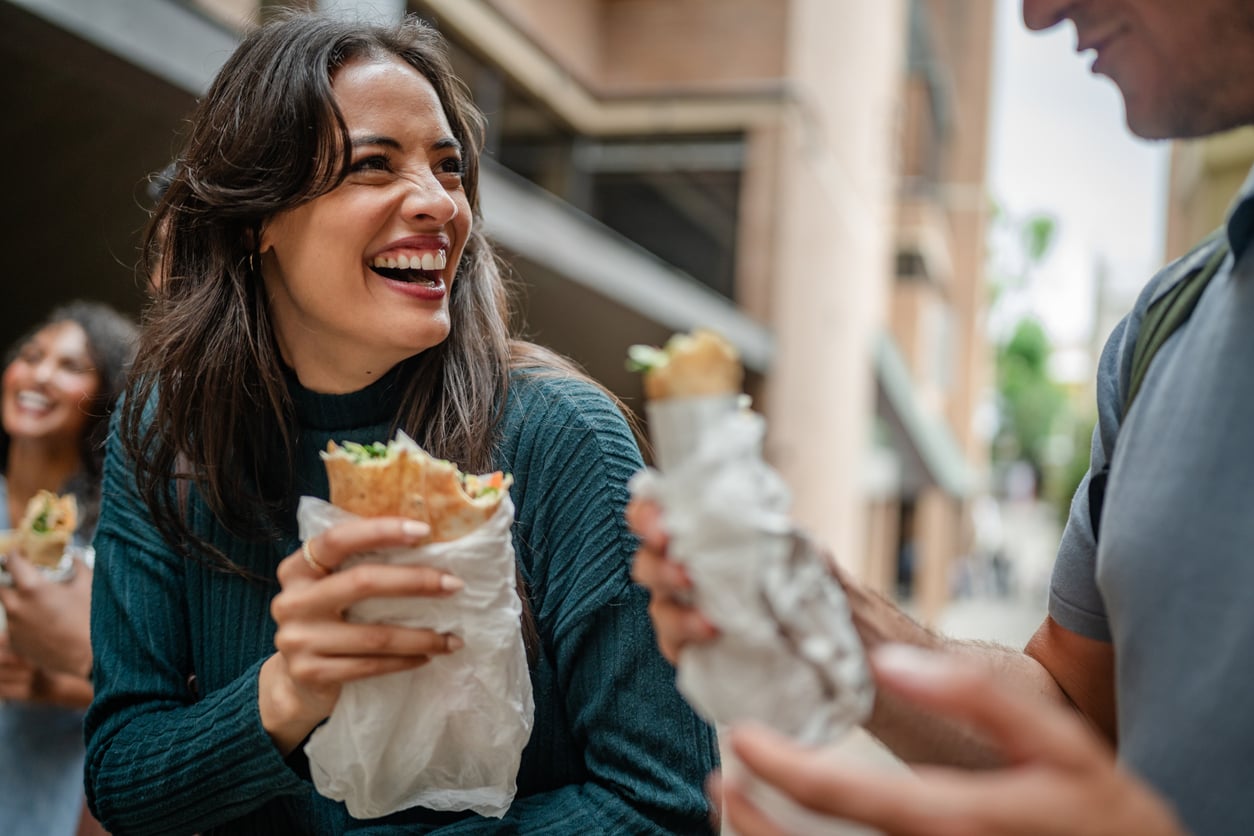 Three people stand outdoors, smiling and holding wrapped sandwiches or burritos; the focus is on a woman in a green sweater laughing with another person, with buildings in the background.