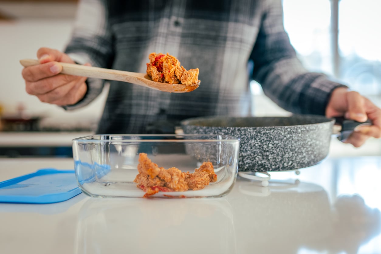 A person in a plaid shirt transfers cooked fried chicken from a pan into a glass container using a wooden spoon.