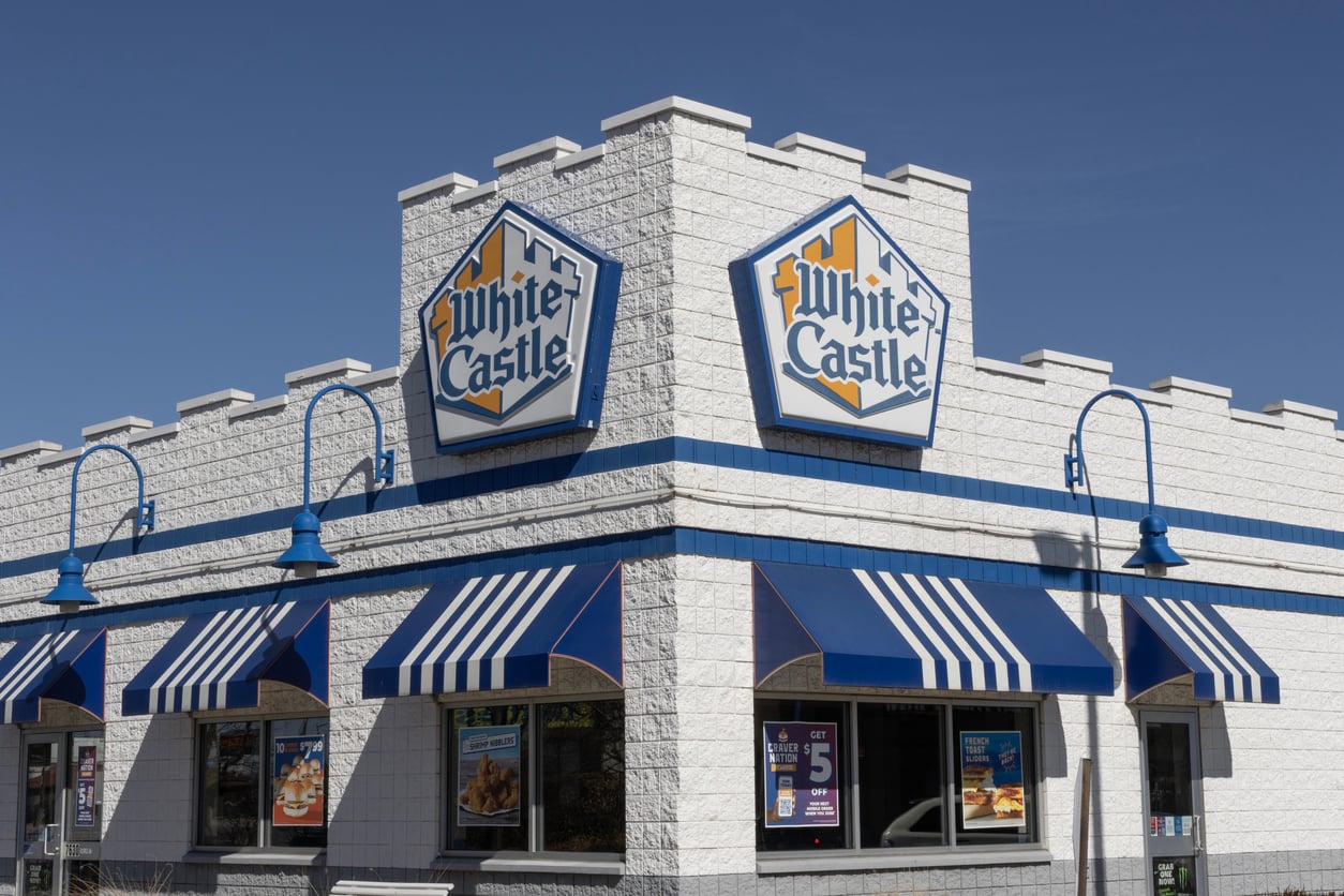 White Castle restaurant exterior with blue and white striped awnings, turret-style architecture, and two large White Castle signs on the corner of the building under a clear blue sky.