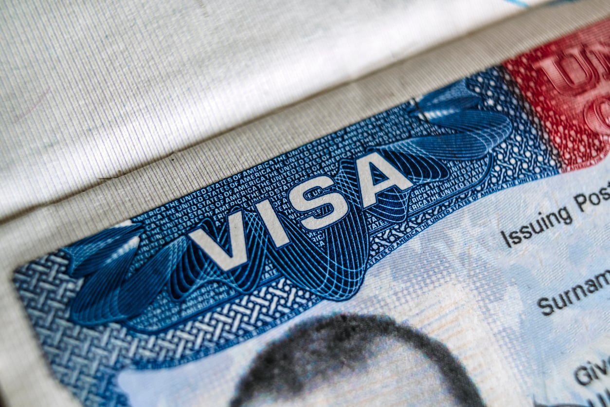 Close-up of a U.S. visa document, showing the word “VISA” in bold blue letters, partial text, and part of a face at the bottom edge—a stark reminder of current travel restrictions and advisories.