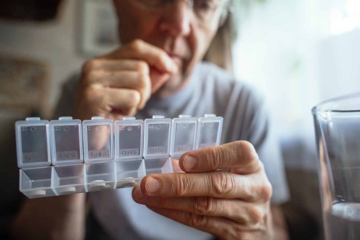 An older person, mindful of their travel habits, holds a weekly pill organizer and prepares to take medication, with a glass of water nearby.