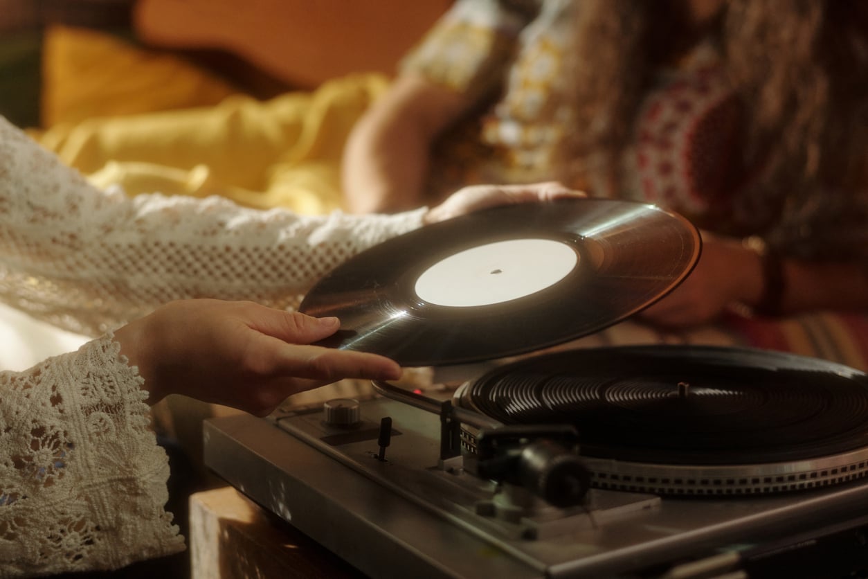 A person in a lacy sleeve holds a vinyl record above a vintage turntable while another person with long hair sits nearby in a patterned shirt. The scene is softly lit and cozy.