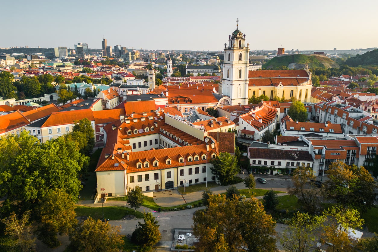Aerial view of Vilnius Old Town with red-roofed buildings, church towers, and green trees under a clear sky. Modern cityscape rises in the background, highlighting why Vilnius has become one of the travel spots boomers are learning to love.