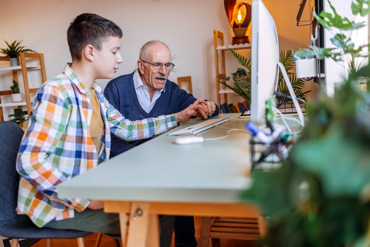 An elderly man and a teenage boy sit at a desk using a computer together. The older man is pointing at the screen while the boy operates the mouse. Indoor home setting with plants and shelves in the background.