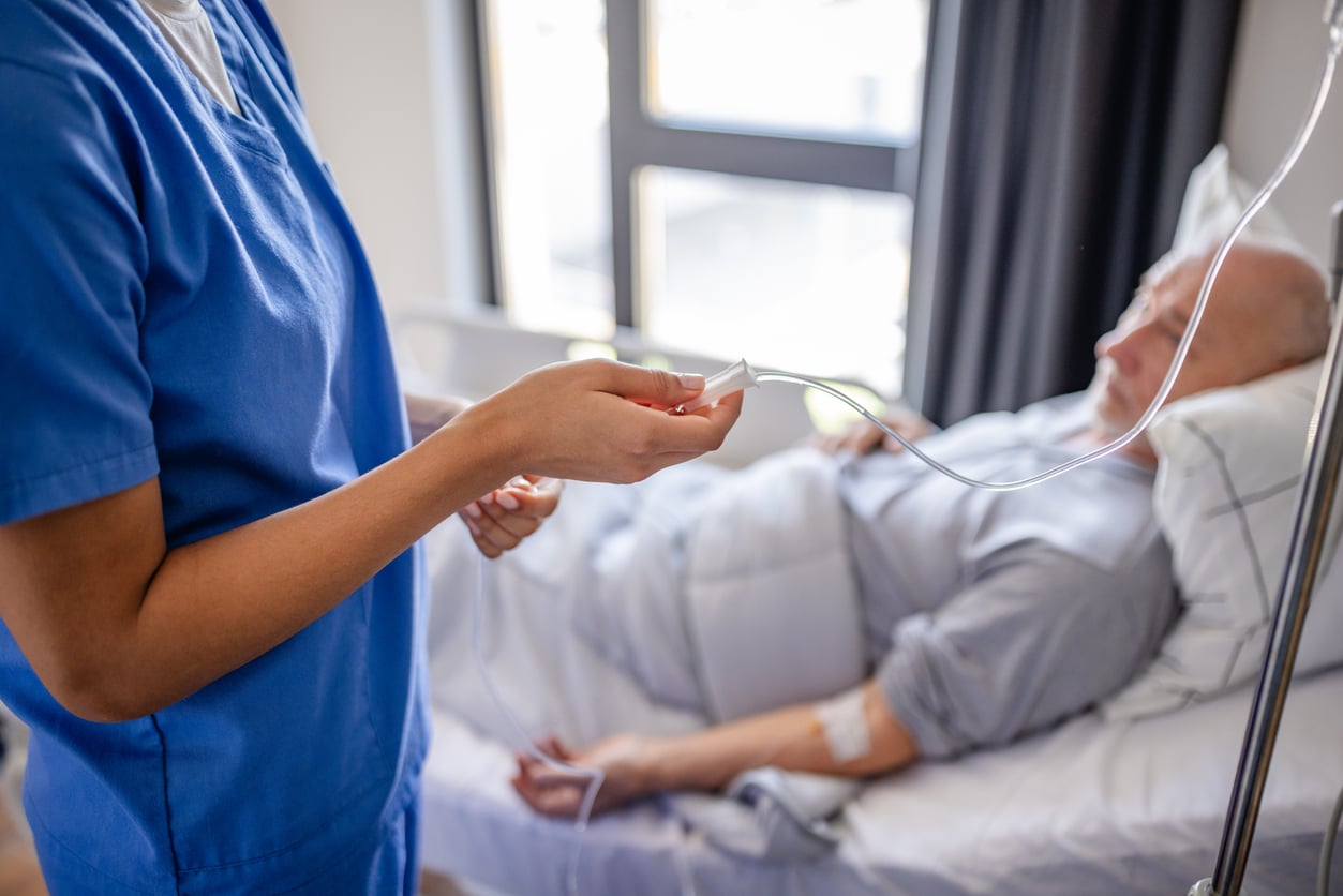 A nurse in blue scrubs adjusts an IV drip for an elderly patient lying in a hospital bed, who appears to be resting with a blanket covering him. Daylight shines through a nearby window.