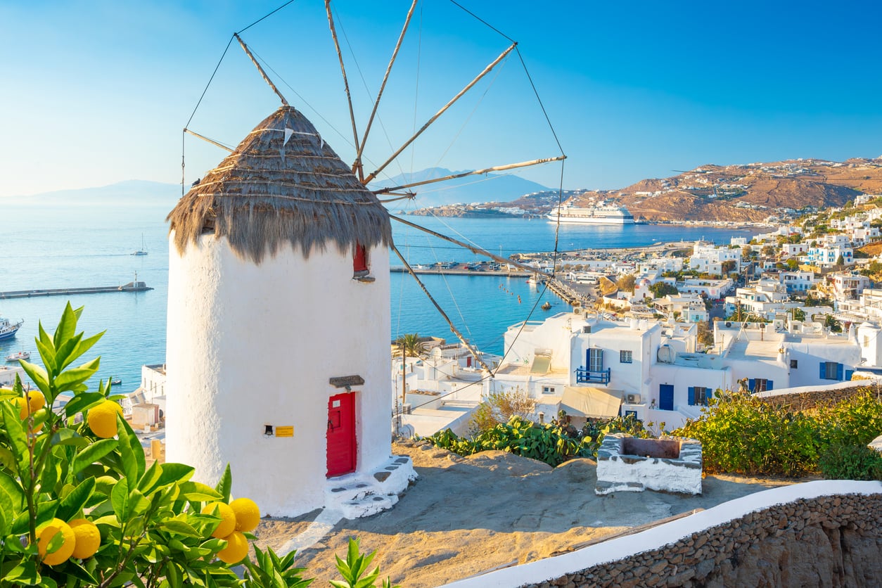 Traditional white windmill with a thatched roof overlooks a coastal town with white buildings and blue accents, bright yellow lemons in the foreground, and a cruise ship in the turquoise sea under a clear blue sky—a picturesque scene from one of the travel destinations boomers are skipping.