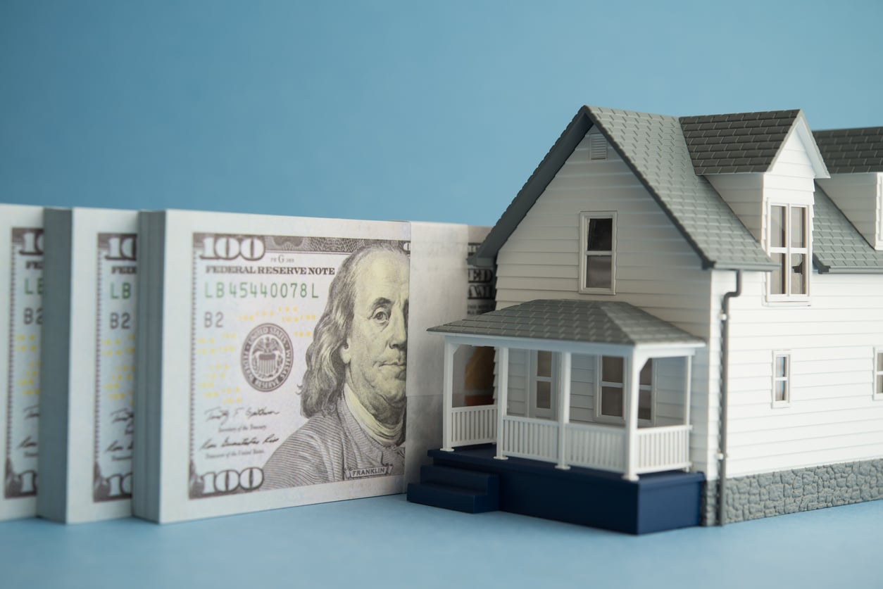 A model house is positioned next to upright stacks of hundred-dollar bills, symbolizing real estate finance or home buying, against a blue background.