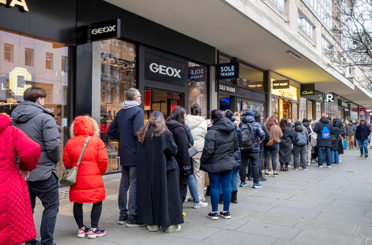 A long line of people wearing winter coats waits outside a row of shops on a city street, including stores like Geox, Sole Trader, and Pop Mart.