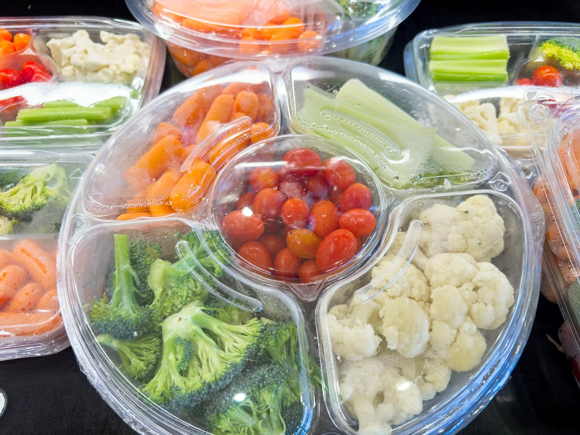 A plastic tray with sections of cherry tomatoes, celery, cauliflower, broccoli, and baby carrots, surrounded by similar vegetable trays, all covered with clear lids.