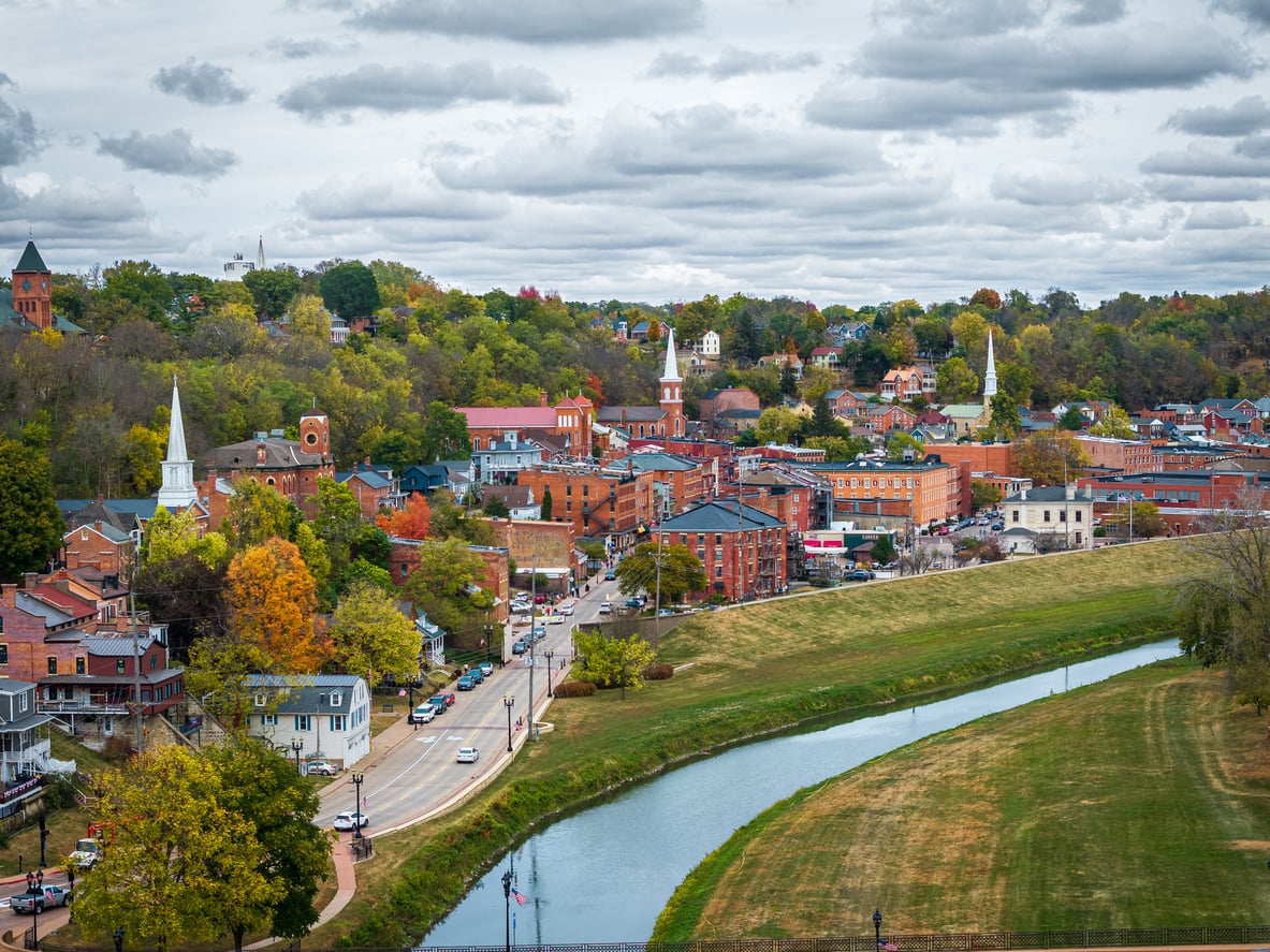 A small town with brick buildings, church steeples, and autumn trees sits beside a winding river under a cloudy sky, embodying the charm of nostalgic travel destinations.