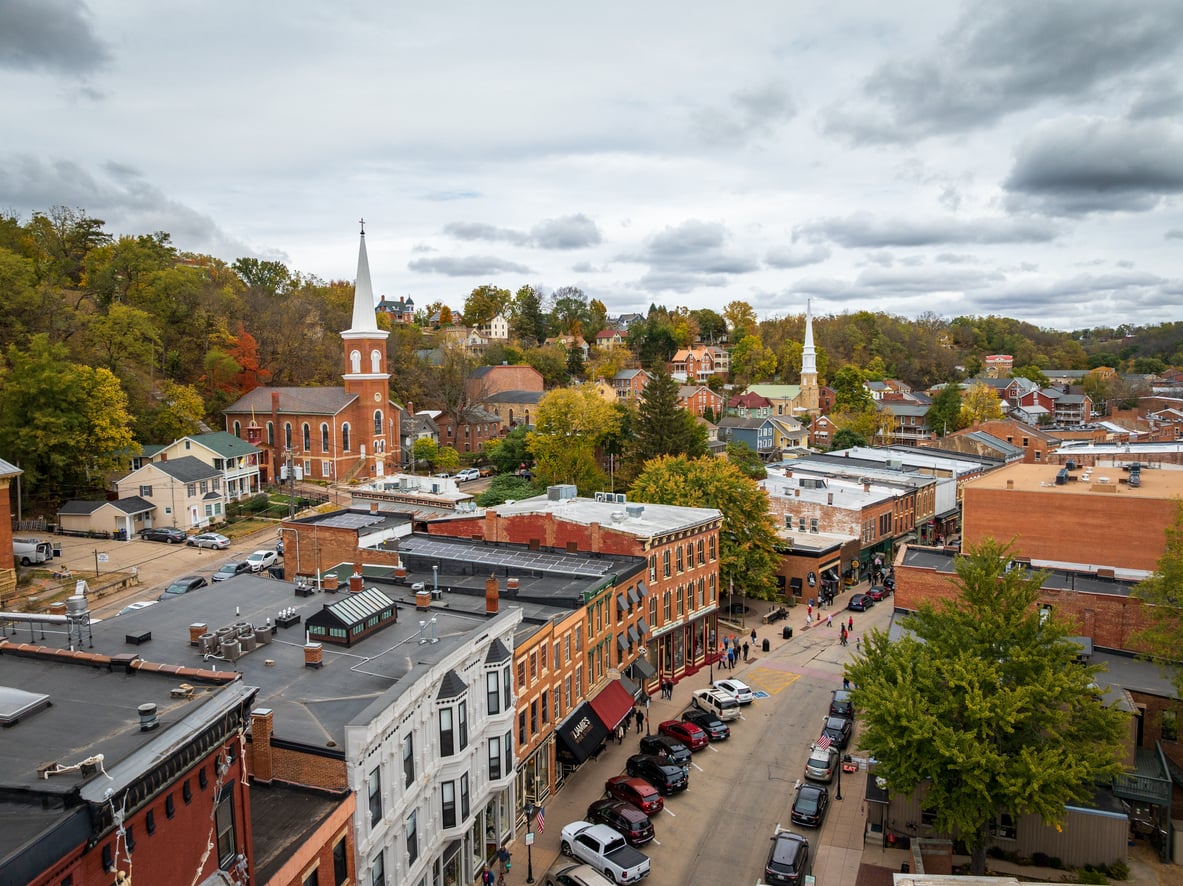 Aerial view of a small town—one of the best fall towns in America—with historic brick buildings, a church with a tall steeple, trees with vibrant autumn foliage, and cars parked along the main street under a cloudy sky.