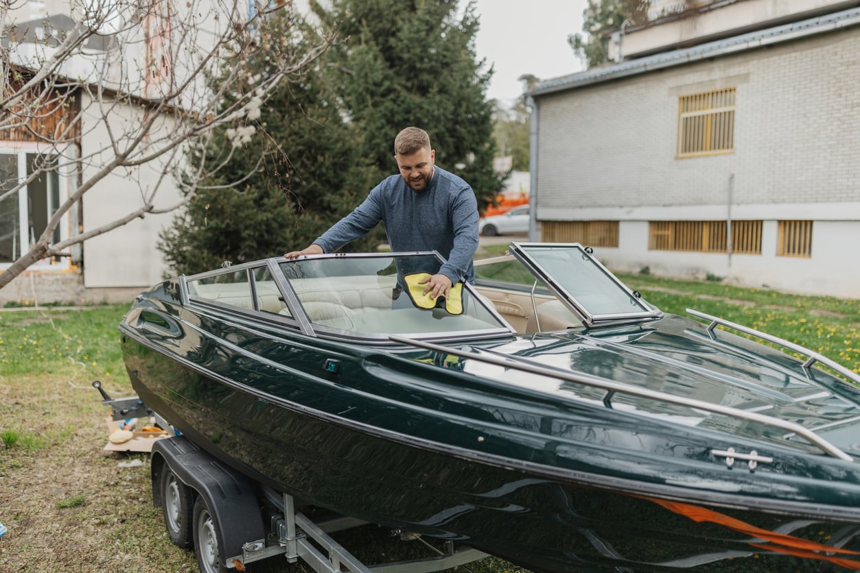A man cleaning the windshield of a dark green boat with a yellow cloth while the boat is on a trailer in a grassy yard.