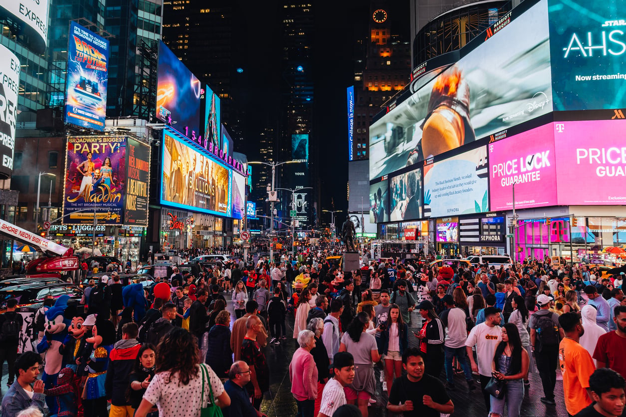 A large crowd fills Times Square at night, surrounded by bright, colorful digital billboards and neon signs. People walk, gather, and take photos amid the lively, bustling city atmosphere.