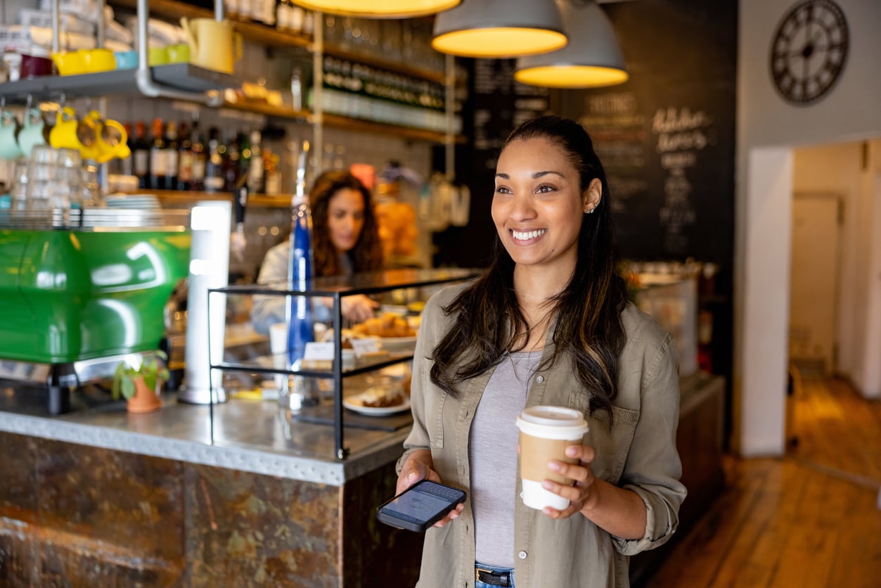 Smiling woman holding a coffee cup and smartphone stands in a cozy café, with pastries and drinks on display and another person working behind the counter. Warm, inviting atmosphere with wooden floors and shelves.
