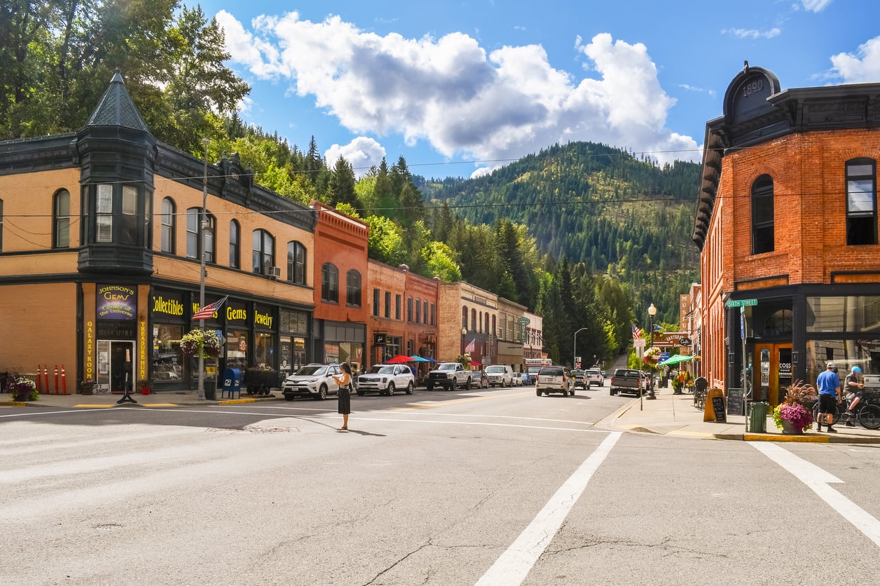 A small mountain town with historic brick buildings, parked cars, and a tree-covered hill in the background under a blue sky with clouds. People are walking along the sidewalks, evoking the charm of nostalgic travel destinations.