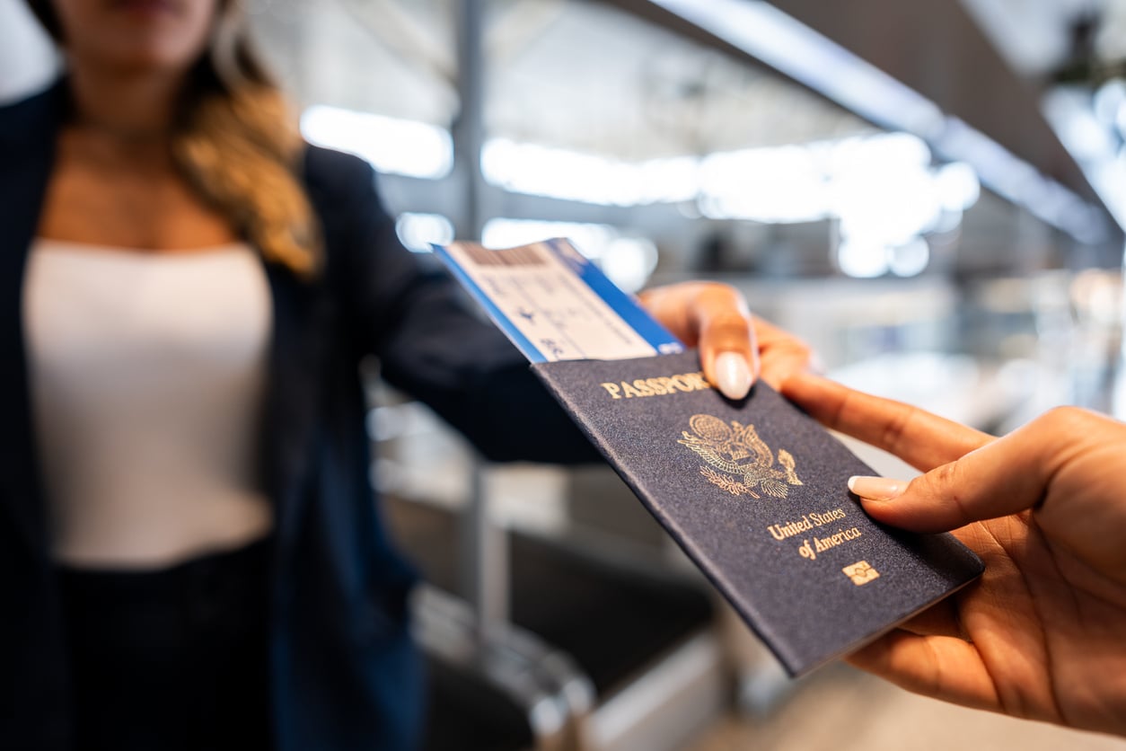 A person hands a U.S. passport and boarding pass to an airline worker at an airport check-in counter, preparing for departure while mindful of current travel restrictions and advisories.