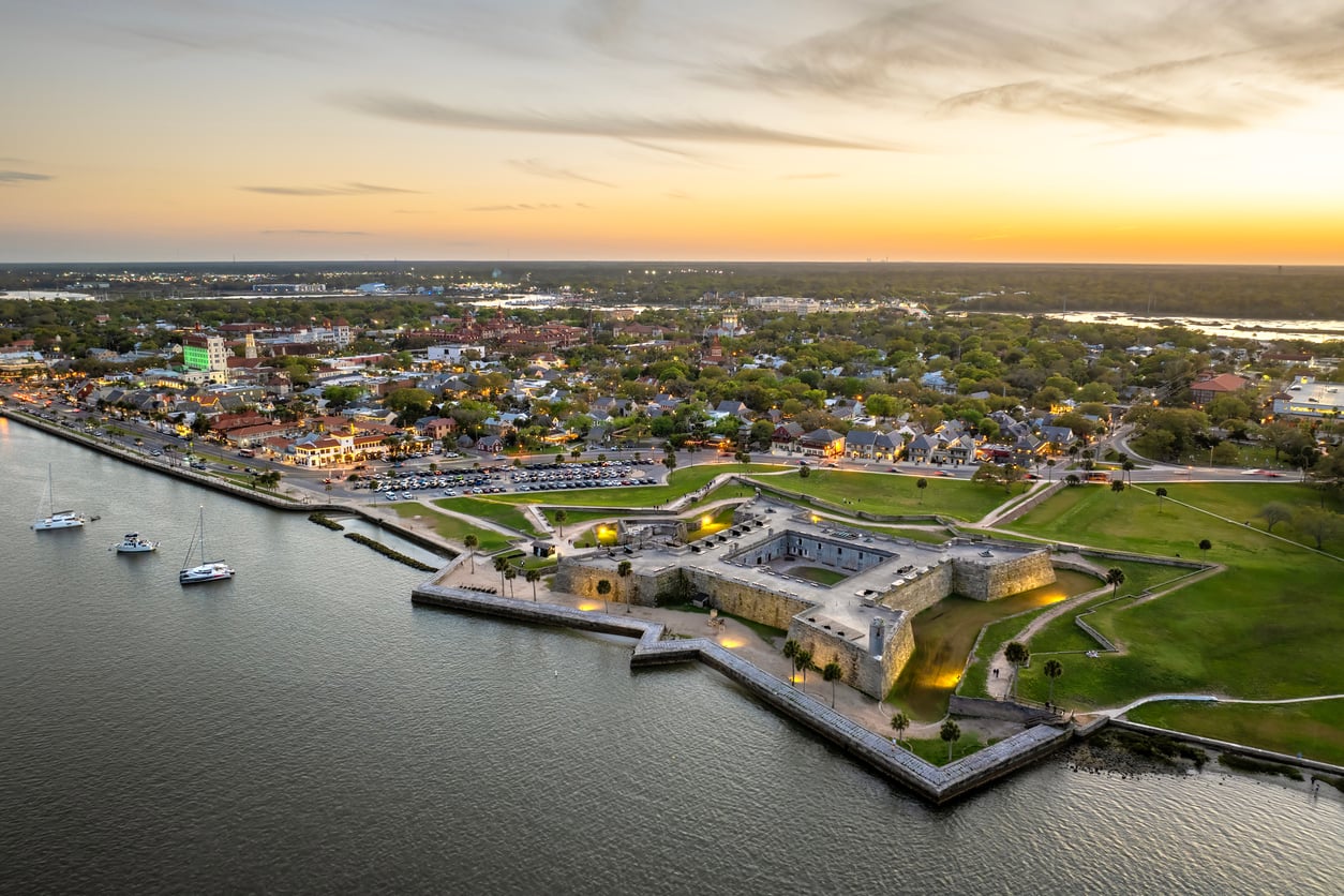 Aerial view of a historic coastal fort by the water at sunset, with boats anchored nearby and a small city in the background under a partly cloudy sky—one of those nostalgic travel destinations that captures the charm of timeless seaside escapes.