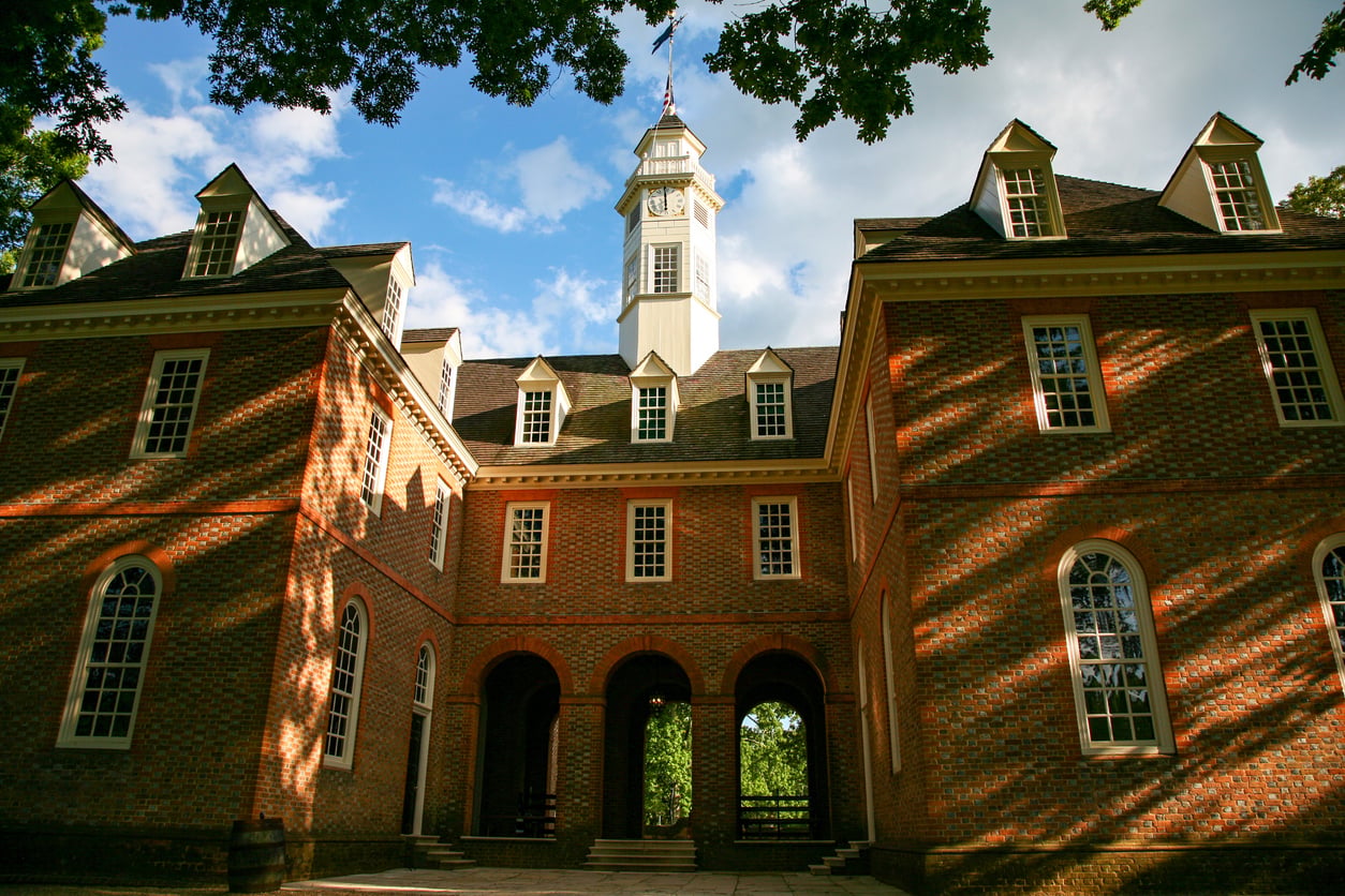 Historic brick building with arched entryways, tall windows, and a central clock tower, pictured under a blue sky with sunlight casting shadows through trees on the facade—a true gem among nostalgic travel destinations.