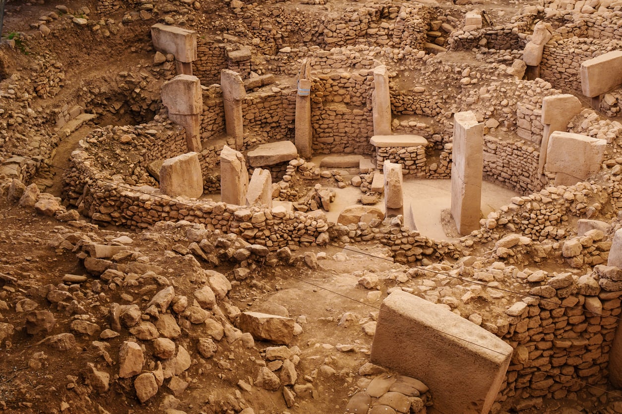 Ancient stone pillars and circular walls are arranged in Göbekli Tepe, an archaeological site with weathered stones and ruins, partially buried in earth and surrounded by rocks.
