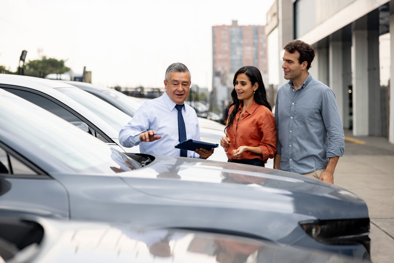 A car salesman shows a couple a row of parked cars outside a dealership. The salesman holds a clipboard and gestures while the couple listens and smiles.