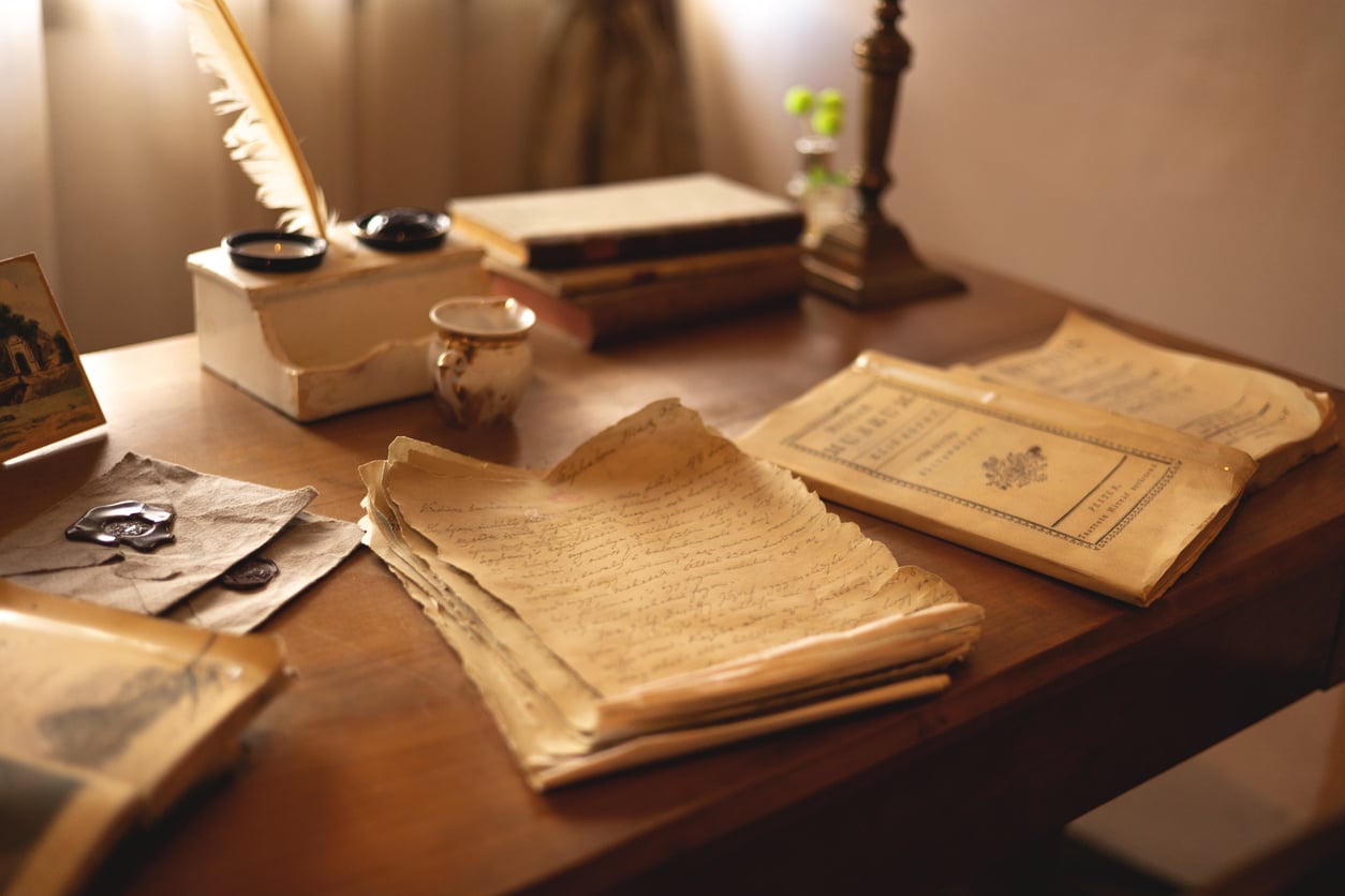 A wooden desk with handwritten papers, vintage books, a feather quill in an inkwell, an old envelope, and a small cup—evoking the charm of things hotels used to offer guests—bathed in warm light from a nearby lamp.