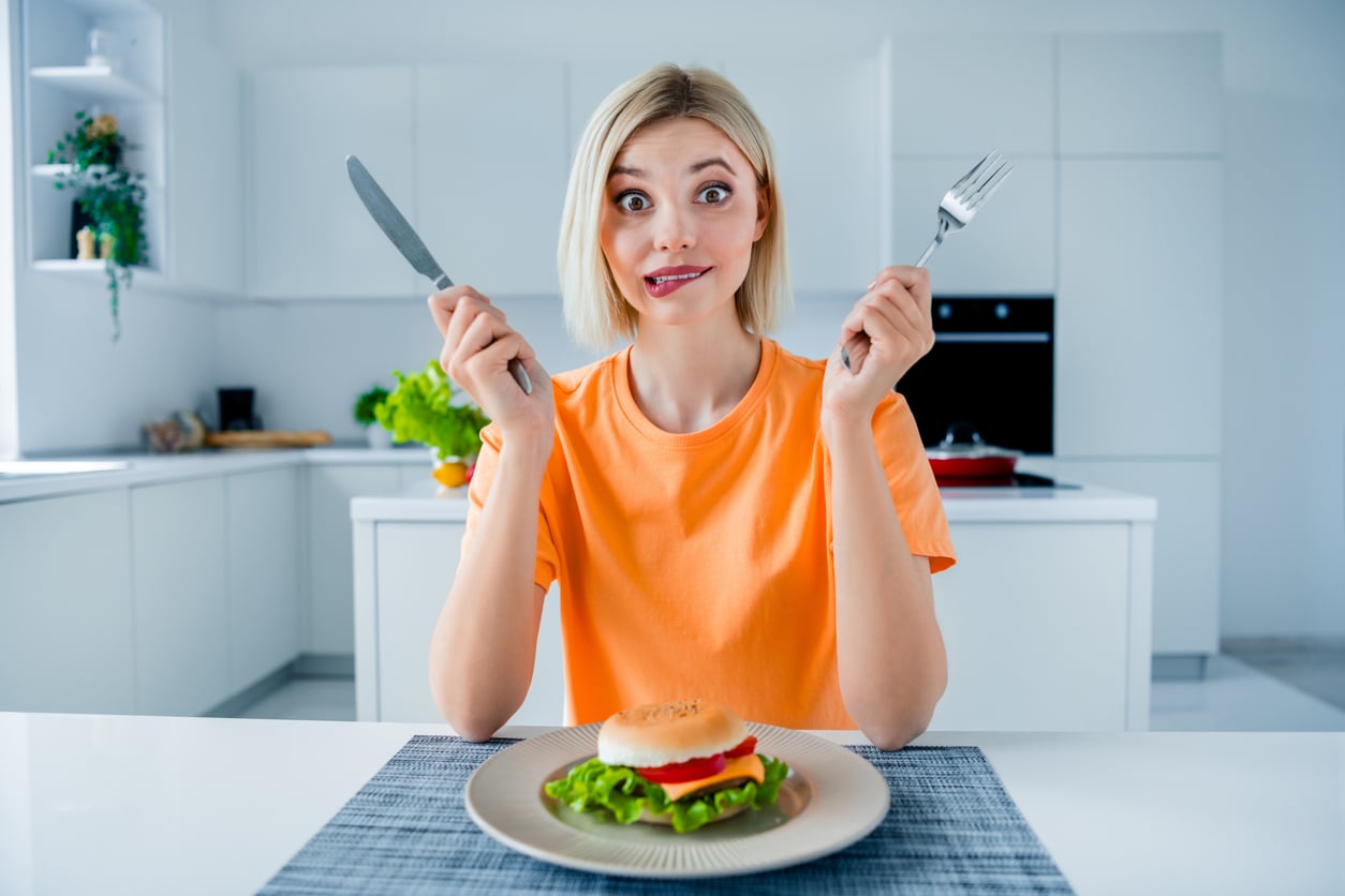 A woman in an orange shirt sits at a table in a modern kitchen, holding a fork and knife, looking excitedly at a plate with a sandwich and lettuce.