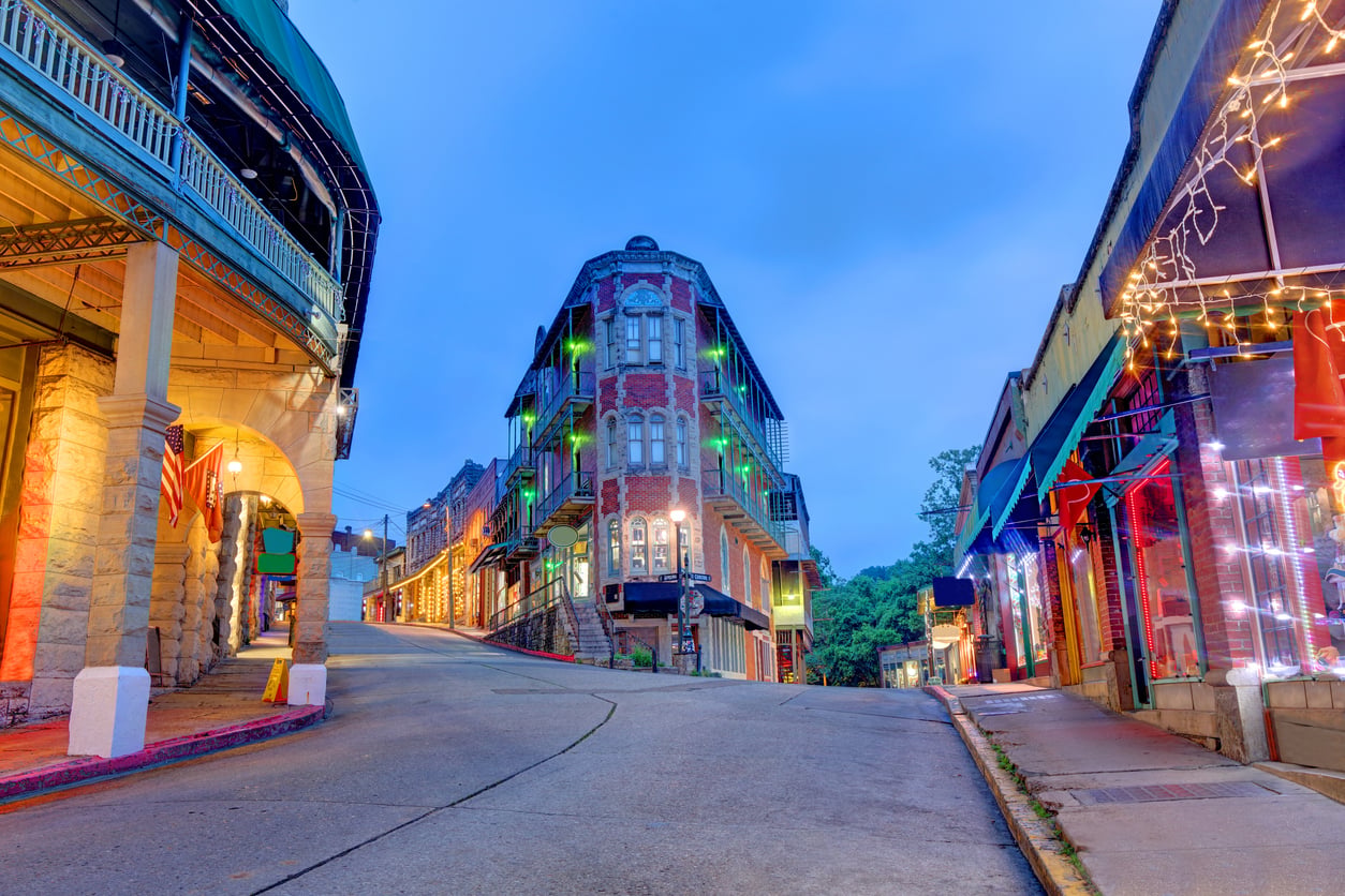 A sloping street in a small town at dusk, lined with colorful buildings and string lights, evokes the charm of nostalgic travel destinations, with a historic brick building standing prominently at the intersection.
